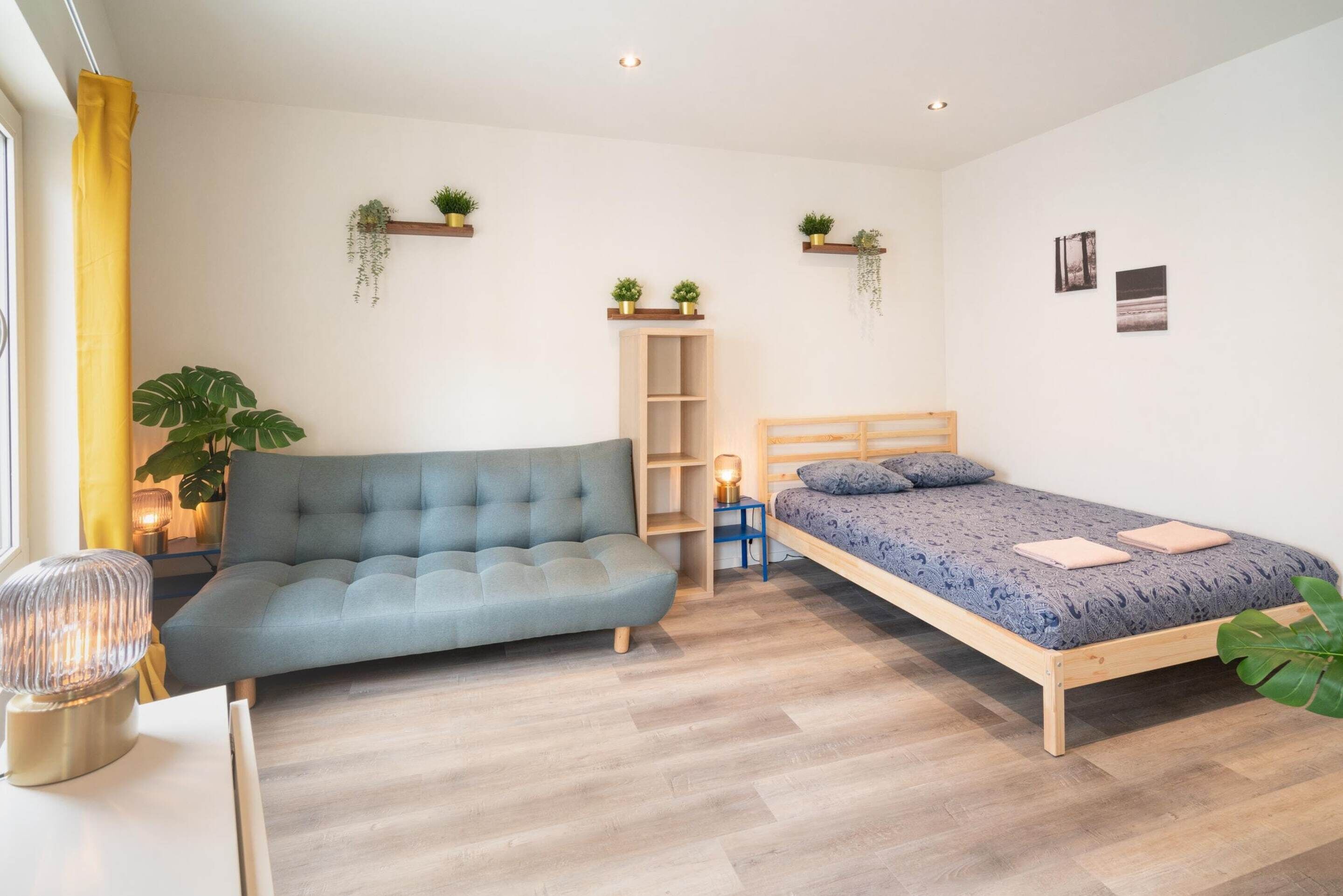A bedroom with a sofa, a bed, a bedside table, a lamp, a shelf with plants, and a wall-mounted shelf.