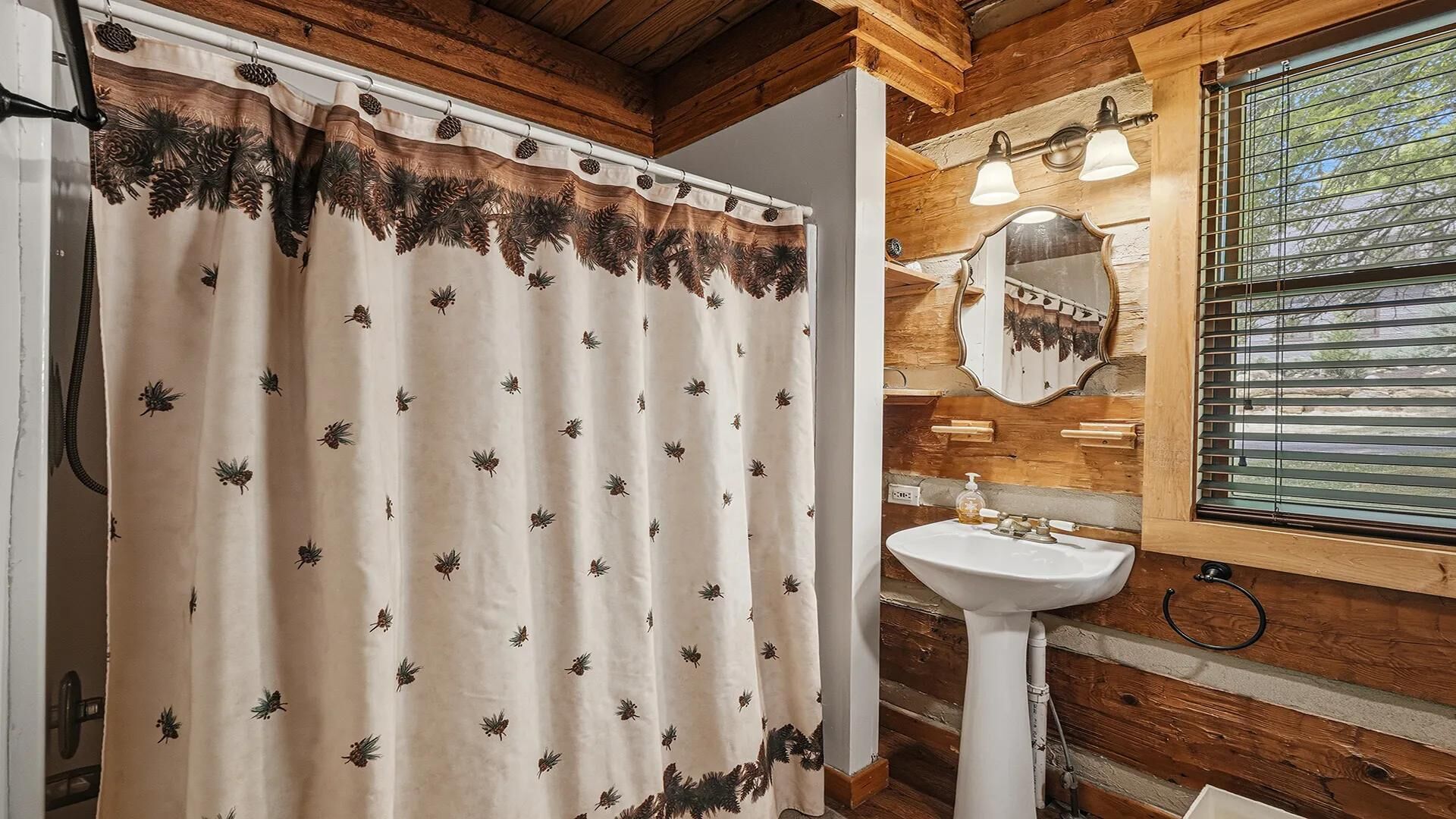 A bathroom with a shower curtain featuring pinecone and pine needle patterns, a white pedestal sink, wooden walls, and a window with blinds.