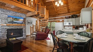 A cozy cabin interior with a stone fireplace, wooden ceiling beams, a dining area, and a kitchen with white appliances. - Appalachian Songbird- Charming 2 bedroom cabin & Pet Friendly (Sevierville)