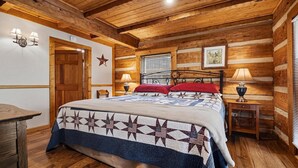 A log cabin-style bedroom with a bed, two bedside tables, a star wall decoration, a window with blinds, and a wooden ceiling. - Appalachian Songbird- Charming 2 bedroom cabin & Pet Friendly (Sevierville)