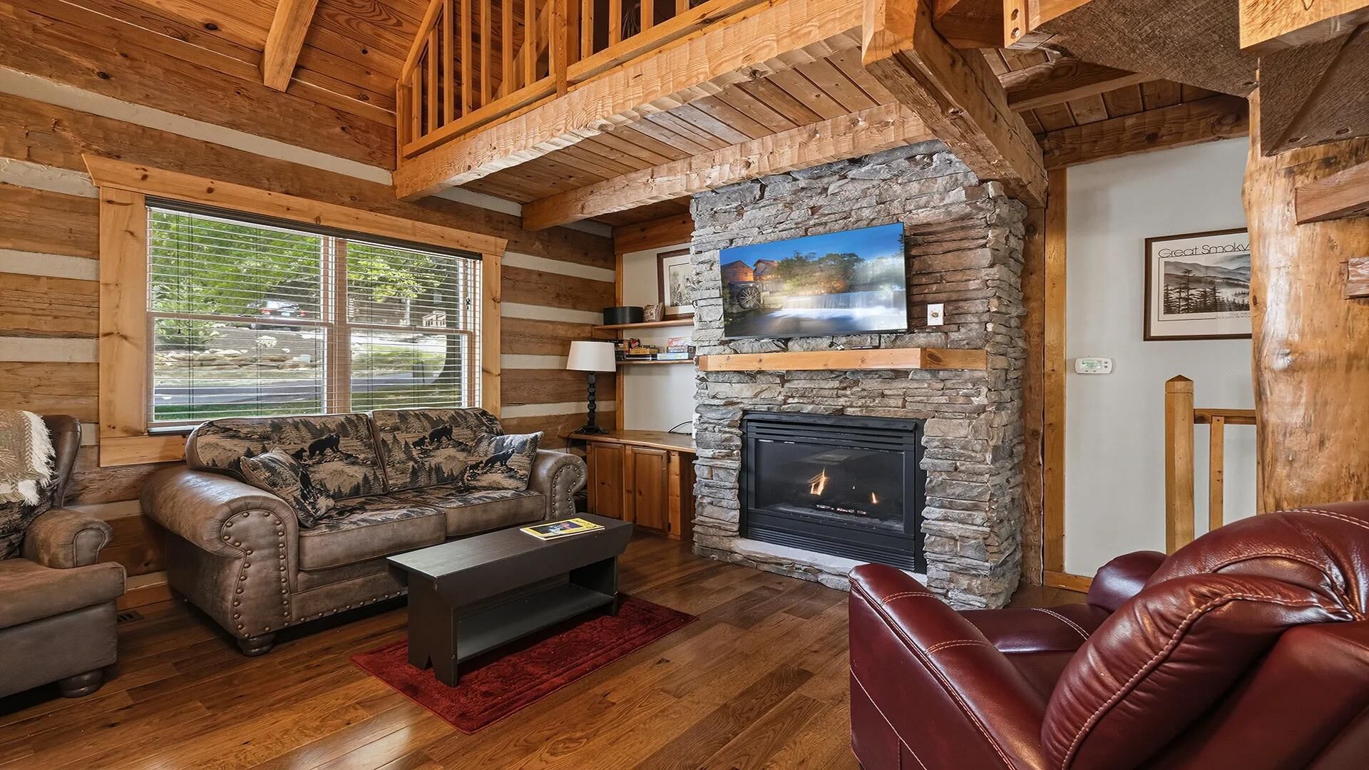A log cabin interior with a stone fireplace, wooden beams, a leather sofa, and a television mounted on the wall.