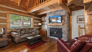 A log cabin interior with a stone fireplace, wooden beams, a leather sofa, and a television mounted on the wall. - Appalachian Songbird- Charming 2 bedroom cabin & Pet Friendly (Sevierville)