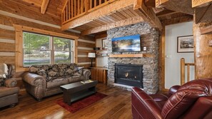 Ein Interieur einer Blockhütte mit Kamin aus Naturstein, Holzbalken, einer Ledersofa und einem an der Wand montierten Fernseher.
