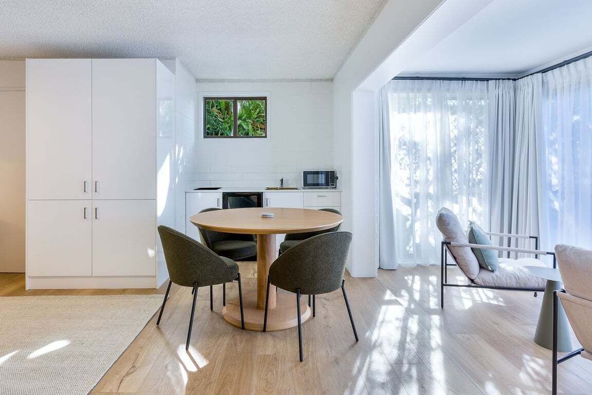 A modern kitchen with white cabinets, a round dining table with chairs, and a view of greenery outside.