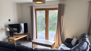 Un salon avec un canapé en cuir noir, une table basse en bois, un téléviseur posé sur un meuble et une porte-fenêtre coulissante donnant sur un espace extérieur.