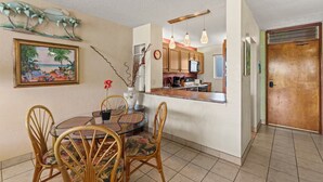 A dining area with a round table and chairs, a kitchen with wooden cabinets, and a door leading to another room. - Oceanfront Menehune Shores #206, Lanai Views, Pool, Steps to Beach, Relax in Kihei (Kihei, Maui)