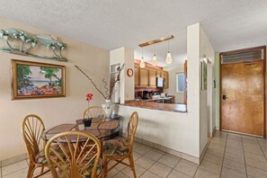 A dining area with a round table and chairs, a kitchen with wooden cabinets, and a door leading to another room. - Oceanfront Menehune Shores #206, Lanai Views, Pool, Steps to Beach, Relax in Kihei (Kihei, Maui)