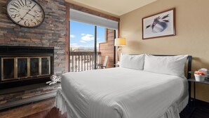 Une chambre d’hôtel avec un grand lit, une cheminée, une horloge, une fenêtre donnant sur un beau paysage et un tableau encadré au mur.