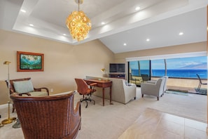 A spacious room with a large window offering a view of the ocean, a comfortable seating area with a wicker chair and sofa, a desk with a chair, and a television.
