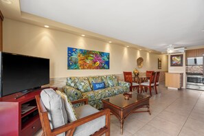 Ein Wohnzimmer mit Flachbildfernseher, einer Couch mit einem Wellen-Kissen, einem Essbereich mit Stühlen und einer Küche mit Kühlschrank.