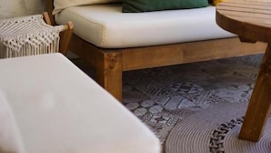 Un banco de madera con cojín, un taburete tejido y una mesa redonda con borde trenzado.
