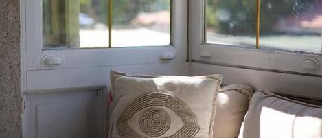 Asiento junto a la ventana con un cojín con un diseño de ojo, y vistas a la vegetación y un edificio.