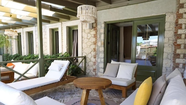 Un área de descanso al aire libre con luz solar, asientos blancos acolchados, una mesa redonda de madera y un muro de piedra de fondo.