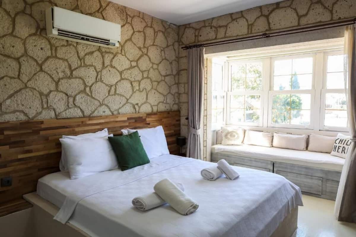 A bedroom with a stone wall, a wooden headboard, a large bed with white bedding, a window seat with cushions, and a view of greenery outside.