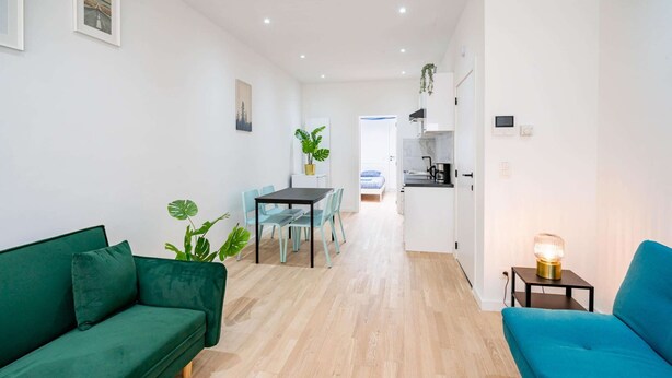 A modern living room with a green sofa, a blue sofa, a dining table with chairs, and a kitchen area in the background.