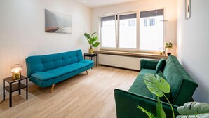 A living room with a blue sofa, a green sofa, a small table with a lamp, and a window with blinds.