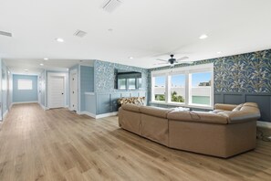 Ein geräumiges Wohnzimmer mit einem großen, beigefarbenen Ecksofa, einem Deckenventilator und Blick durch ein großes Fenster.