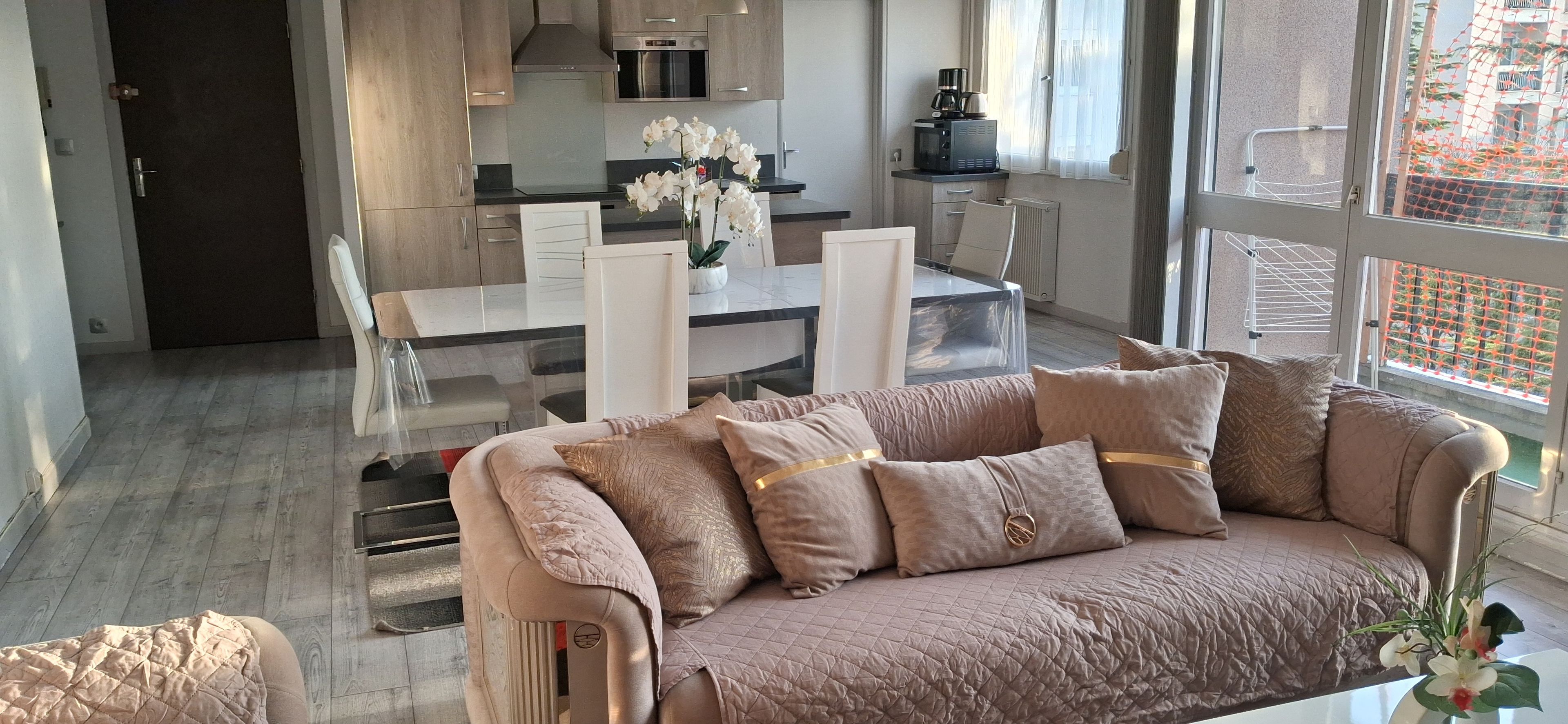 A modern living room with a sofa, a dining table, and chairs. There is a kitchen area with appliances and a window with a view.