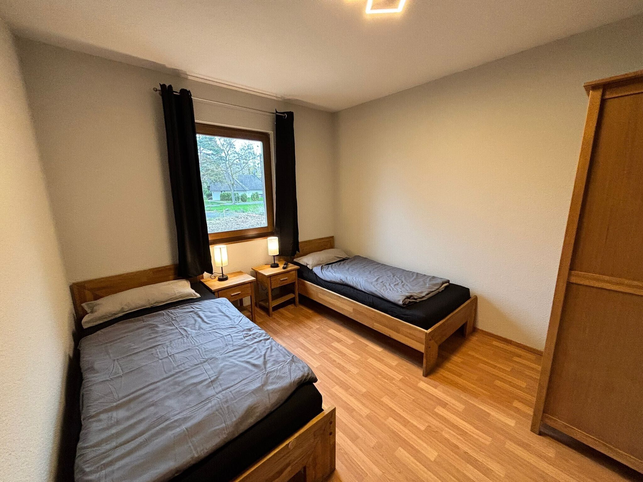 Una habitación pequeña y limpia con dos camas individuales, un armario de madera, una ventana con cortinas y una mesita de noche con lámpara.