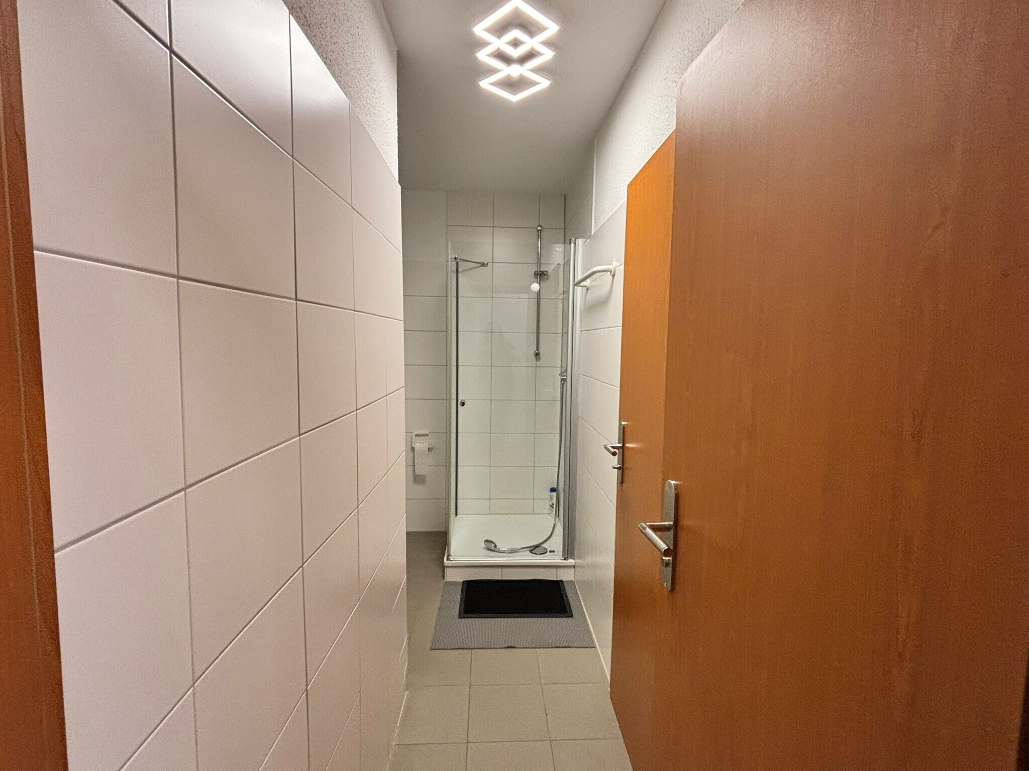 Un couloir étroit aux murs carrelés, une porte en bois et une cabine de douche en verre.
