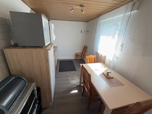 Een kleine keuken met een magnetron, fornuis en houten kastjes. Er is een eettafel met stoelen en een raam dat zorgt voor natuurlijk licht.