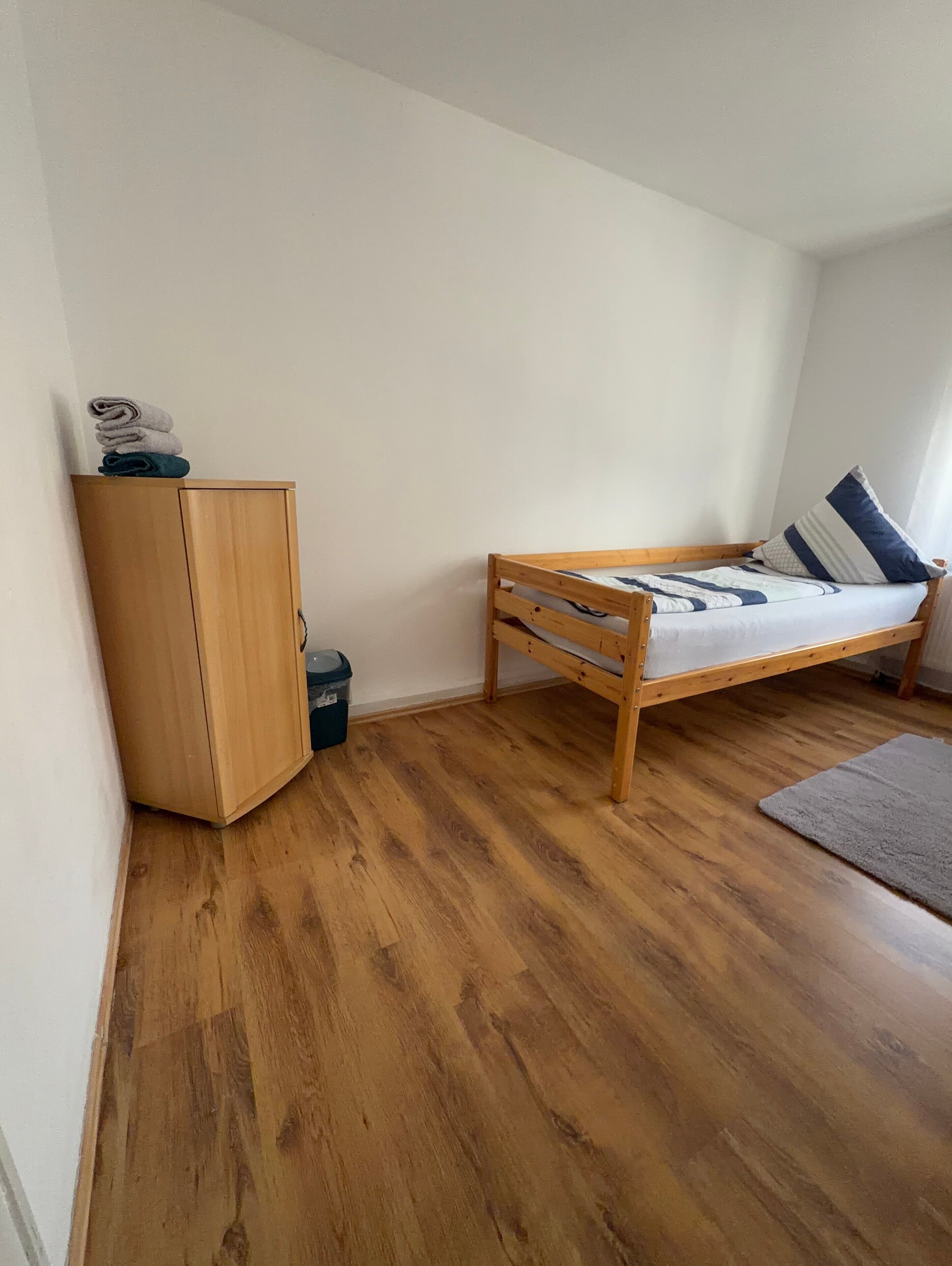 A small, single-room apartment with a wooden wardrobe, a bed with a blue and white striped pillow, and a grey rug on the wooden floor.