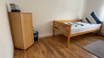 A small, single-room apartment with a wooden wardrobe, a bed with a blue and white striped pillow, and a grey rug on the wooden floor.