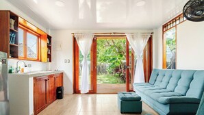 A modern kitchen with wooden cabinets, a built-in sink, and a refrigerator. There is a blue sofa and a small ottoman in a living area with a sliding glass door leading to a garden.