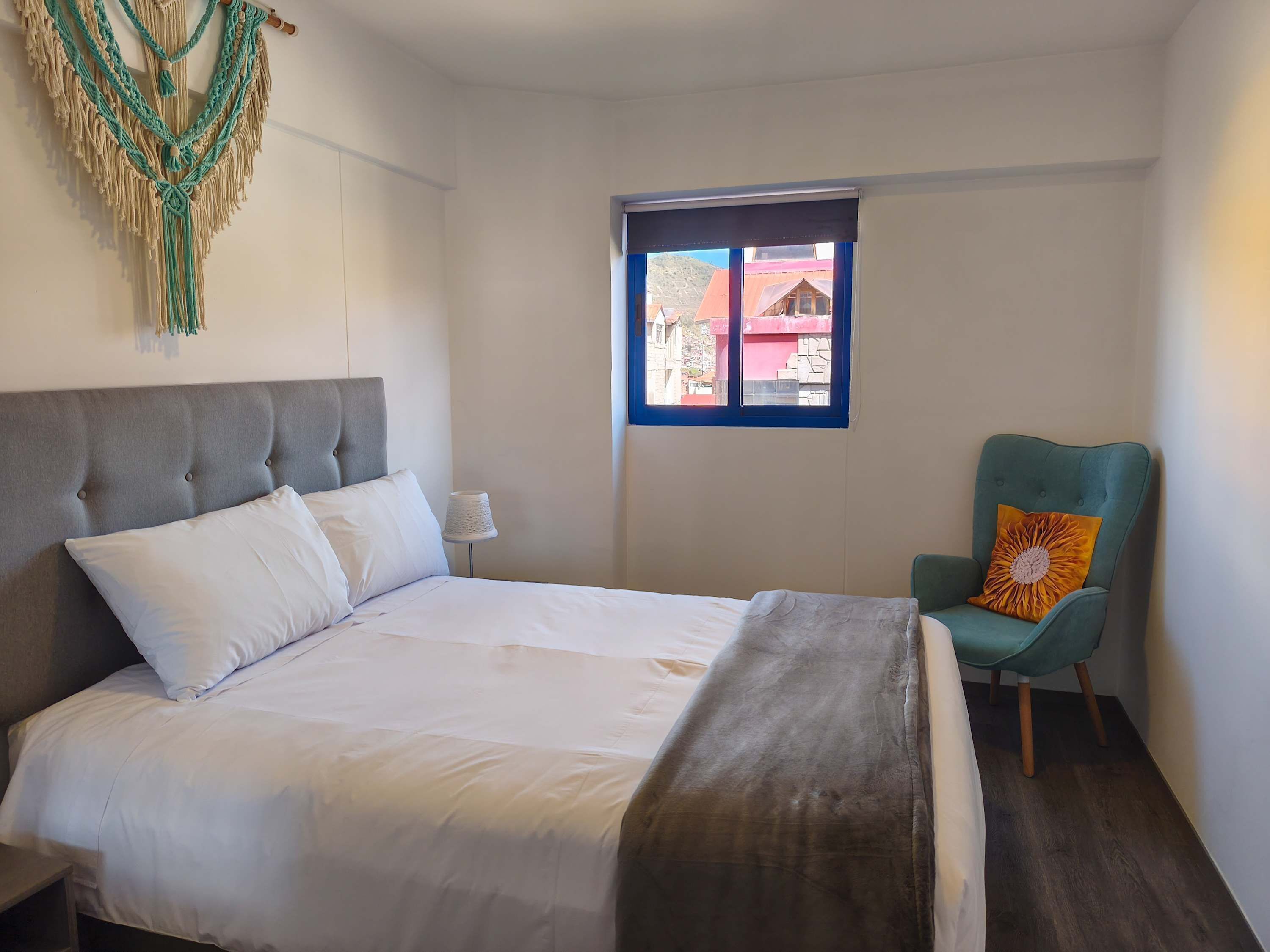 A neatly made bed with white linens, a grey upholstered headboard, a teal armchair with a yellow pillow, and a macrame wall hanging.