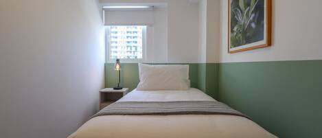 Habitación de hotel estrecha con cama, mesita de noche, lámpara, ventana y un cuadro en la pared.