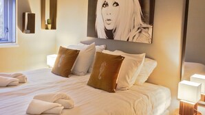 Een opgemaakt bed met kussens, een nachtkastje met een lamp en een kunstwerk aan de muur.