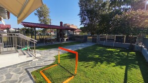 A patio area with a stone floor, a grassy lawn, a fenced play area, and a covered seating area with a red roof.