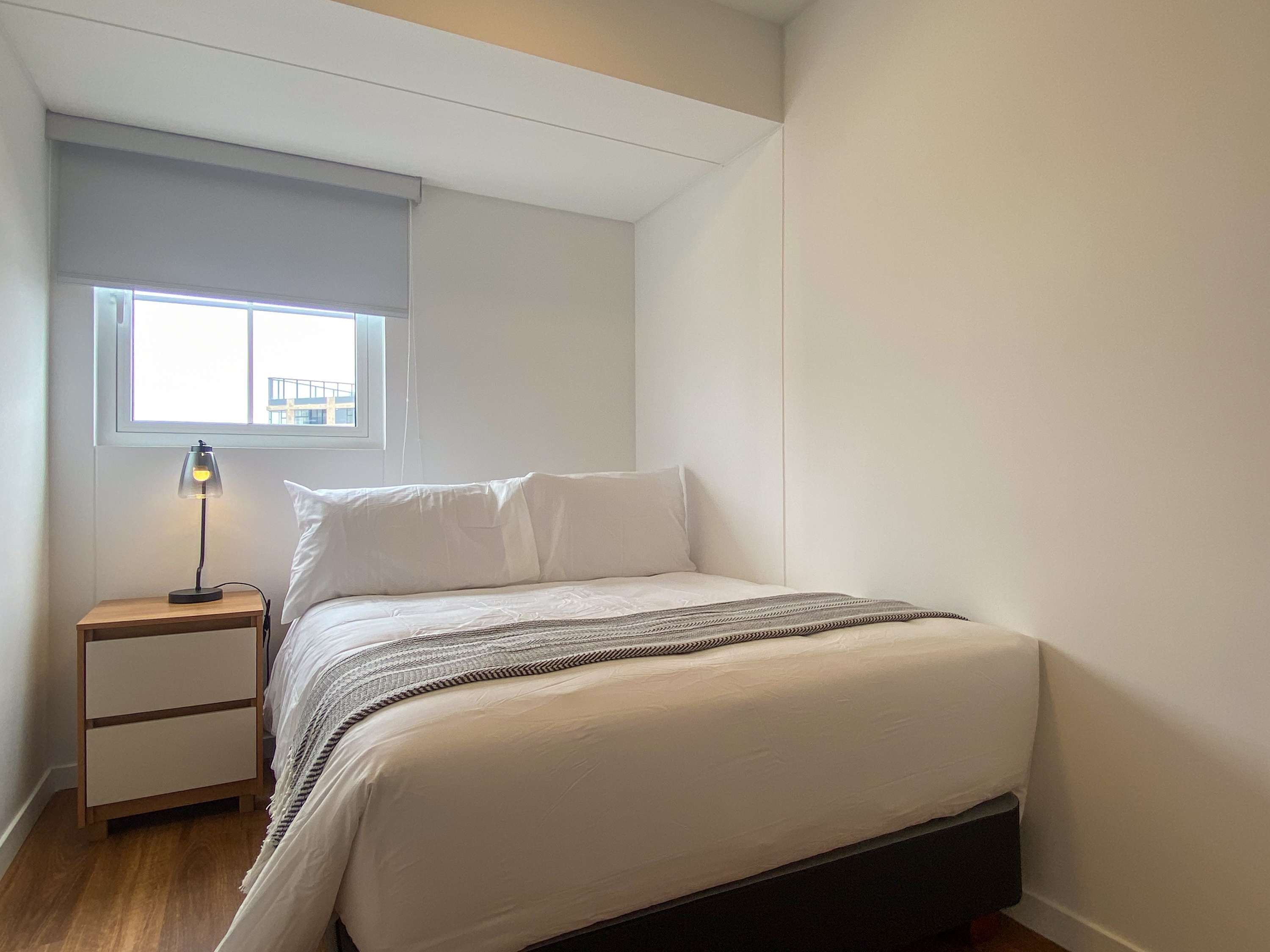 A bedroom with a bed, a nightstand, a lamp, a window, and a view of buildings outside.