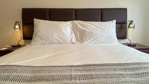 Ein ordentlich bezogenes Bett mit dunklem Kopfteil, zwei Nachttischlampen und einer gemusterten Tagesdecke am Fußende.