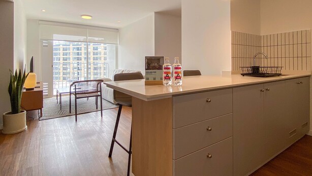 A modern kitchen with a central island, a dining area with a chair, a potted plant, and a view of a cityscape through a large window. - w Upscale 2BR w Balcony View in Miraflores (Lima)
