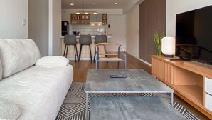 A modern living room with a sofa, a coffee table, and a TV. There is a kitchen in the background with bar stools and a dining table. - w Upscale 2BR w Balcony View in Miraflores (Lima)