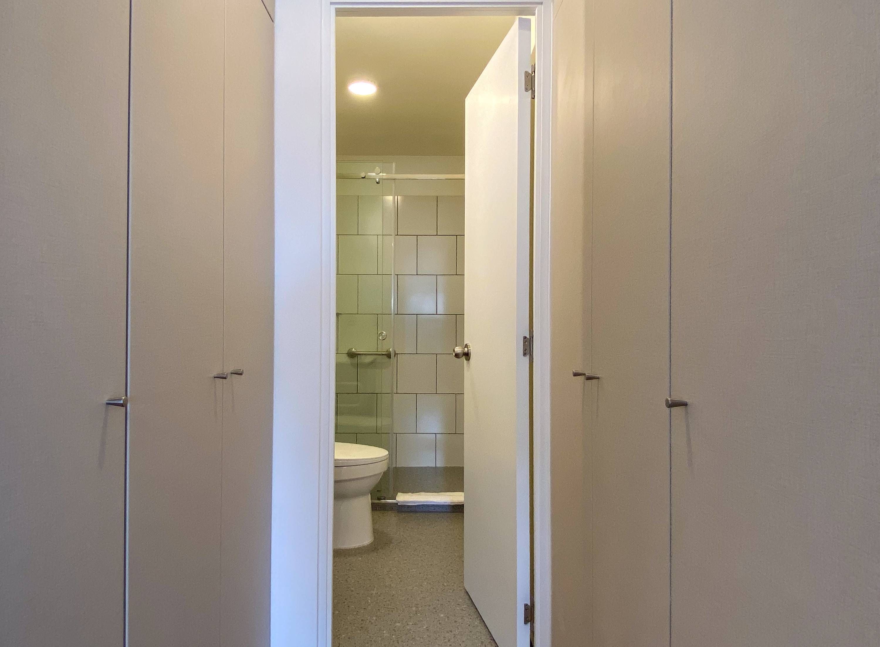 Een smalle gang die uitkomt bij een badkamer met een toilet, een douchegedeelte en een douchecabine met glazen wanden.