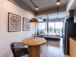 Ein modernes Hotelzimmer mit einem runden Holztisch, einem schwarzen Metallstuhl, zwei gerahmten Bildern an der Wand, einem Bett mit Blick und einem schwarzen Kühlschrank.
