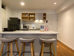 Eine moderne Küche mit weißer Arbeitsplatte, drei Barhocker, einer Kaffeemaschine und einem Kühlschrank.