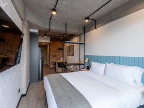 Ein modernes Hotelzimmer mit einem großen Bett, einem Schreibtisch mit Stuhl, einem kleinen Tisch und einer Küchenzeile im Hintergrund.