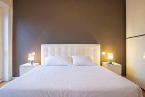 A neatly made bed with white bedding, two bedside tables with lamps, and a plain wall. - Bright Ambrose I Elegant 3 Bedroom Apartment With Balconies in Milan (Milan)