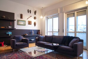 A modern living room with a sofa, armchair, and coffee table, decorated with wall art and a patterned rug. - Bright Ambrose I Elegant 3 Bedroom Apartment With Balconies in Milan (Milan)
