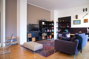 A modern living room with a sofa, ottoman, bookshelf, television, and a coffee table. - Bright Ambrose I Elegant 3 Bedroom Apartment With Balconies in Milan (Milan)