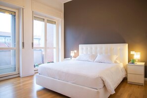 A bedroom with a large bed, a nightstand with a lamp, and a window with blinds. - Bright Ambrose I Elegant 3 Bedroom Apartment With Balconies in Milan (Milan)