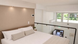 A hotel room with a bed, a small bedside table with a plant, a window with a view of greenery, and a balcony railing. - Athens Mets Loft - Cozy Urban Gem Park Views (Athens)
