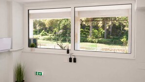 A room with two large windows, a potted plant, a wall-mounted light switch, and a small shelf with a plant. - Athens Mets Loft - Cozy Urban Gem Park Views (Athens)