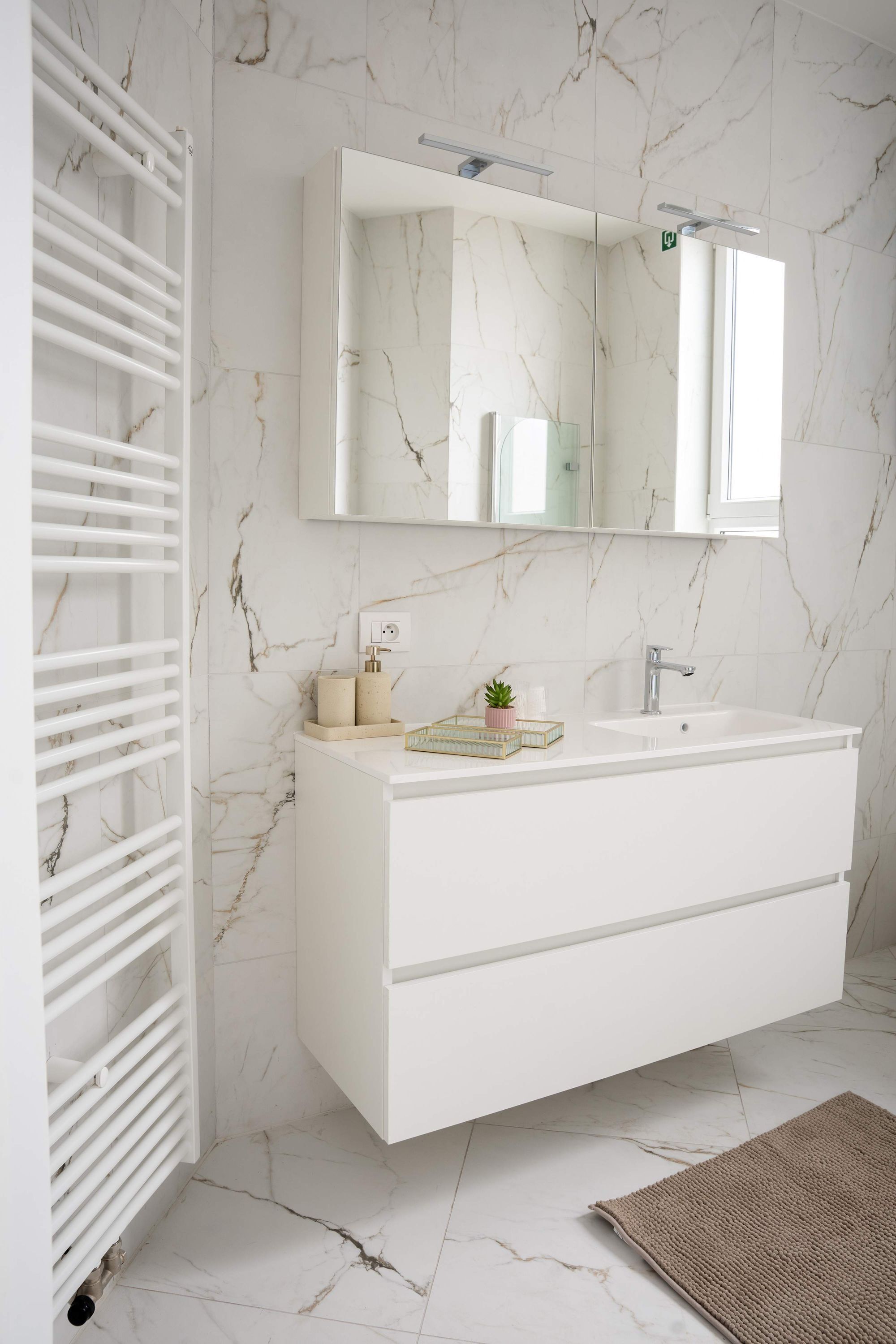 Une salle de bain avec un meuble-lavabo blanc, des murs en marbre, un grand miroir et un porte-serviettes.