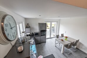 A modern kitchen with a large wall clock, a dining area with a table and chairs, and a view through the glass doors to the outside. - Holiday Home 'Kronseeliebe' with Private Terrace, Garden, and Wi-Fi (Lychen)
