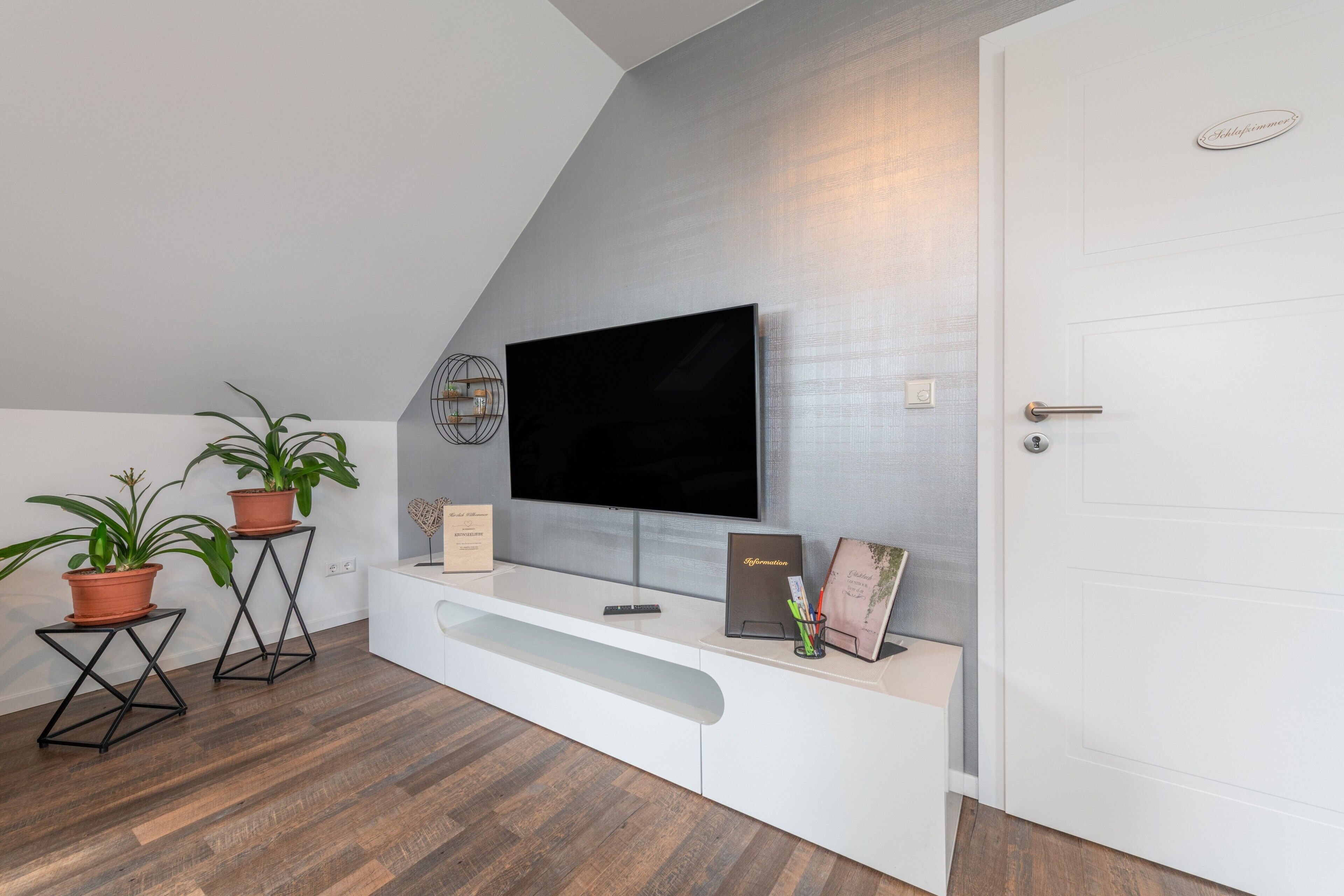 Una sala moderna con un televisor de pantalla plana montado en la pared, un mueble blanco para entretenimiento con un control remoto, una planta en maceta y una puerta.