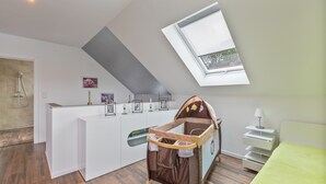 Ein moderner, gut beleuchteter Raum mit schrägem Dach, einem kleinen Fenster, einem Babybett, einer Duschkabine und einem weißen Empfangstresen.